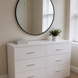 Frost white 6-drawer chest of drawers with slim silver bar handles, styled beneath a round black-framed mirror with books and a potted plant