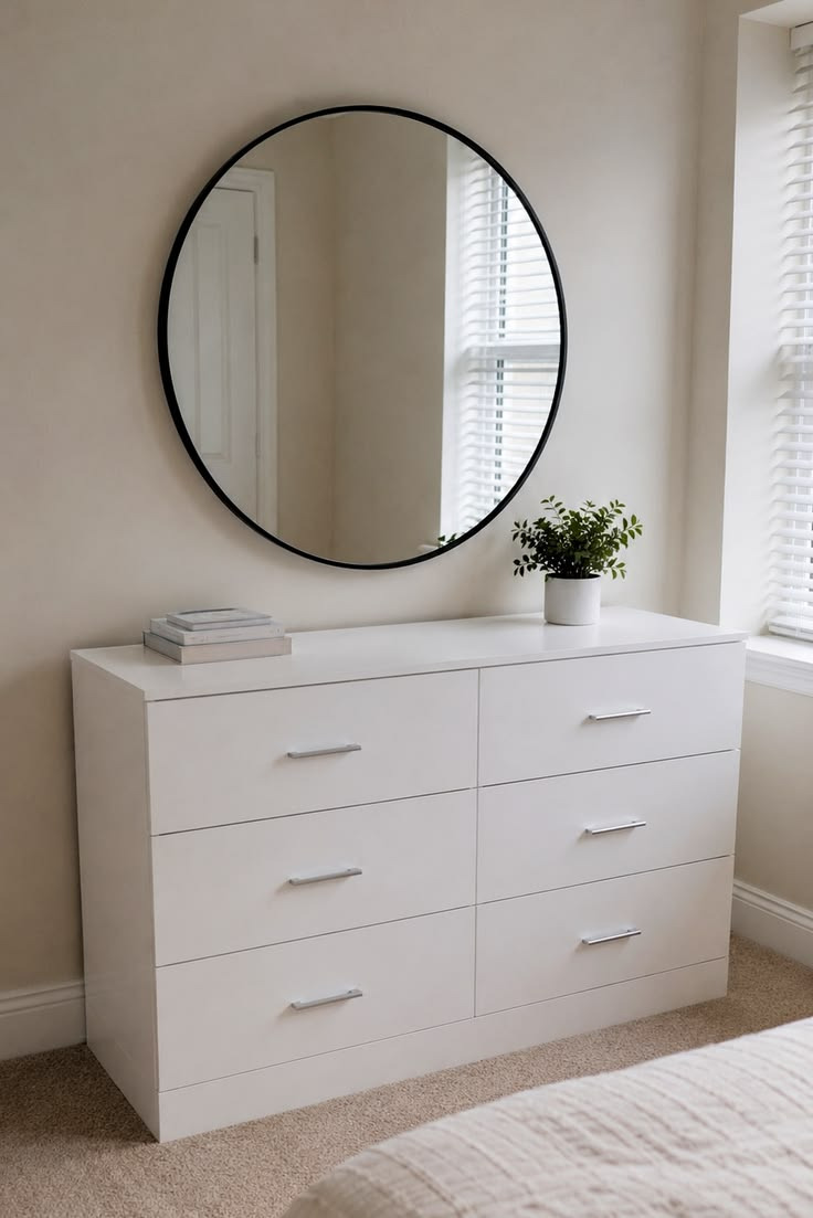 Frost white 6-drawer chest of drawers with slim silver bar handles, styled beneath a round black-framed mirror with books and a potted plant